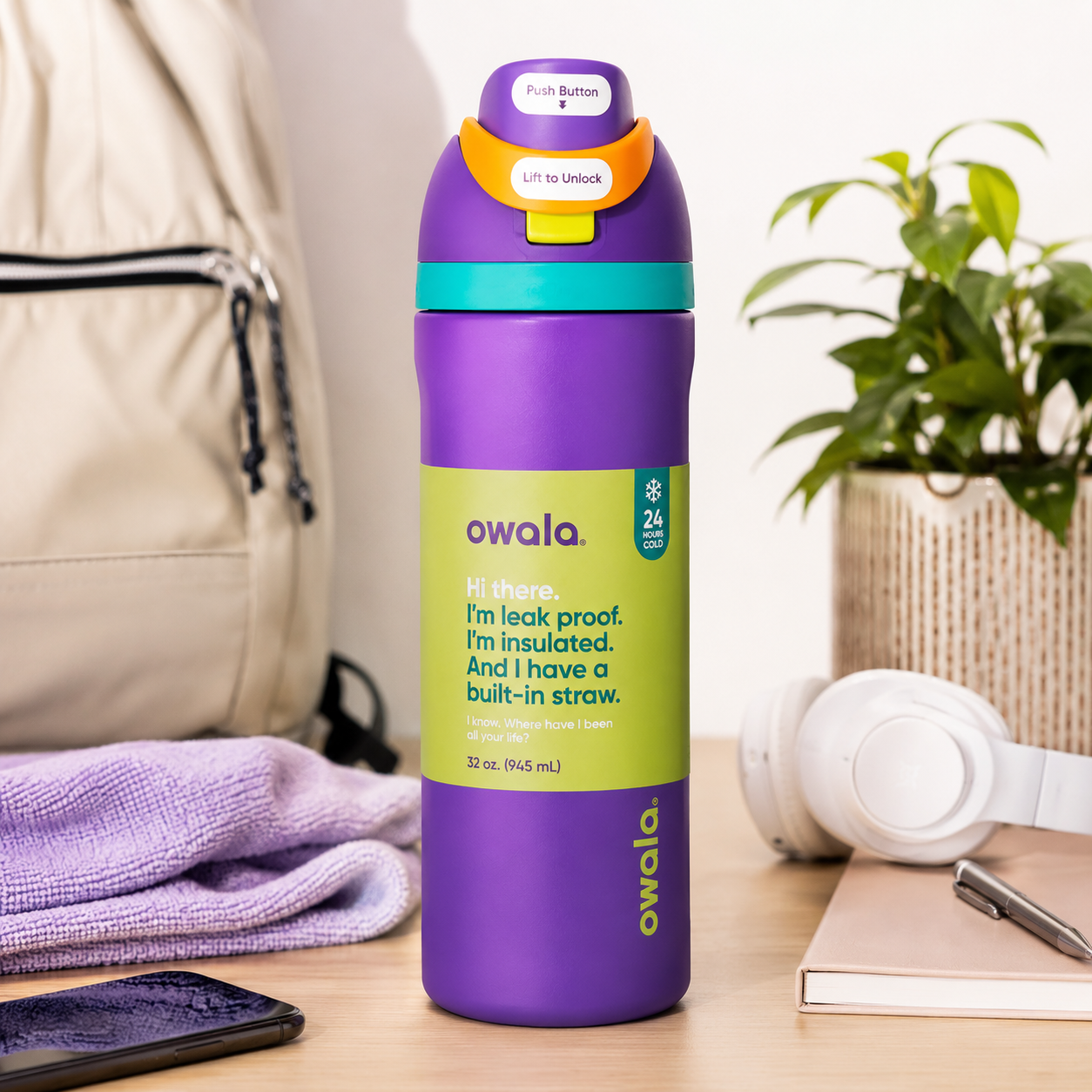 A purple Owala insulated water bottle with a bright green label and a teal-and-orange flip-top lid, standing on a wooden table in a styled room setting with a phone, towel, headphones, notebook, and plant in the background.