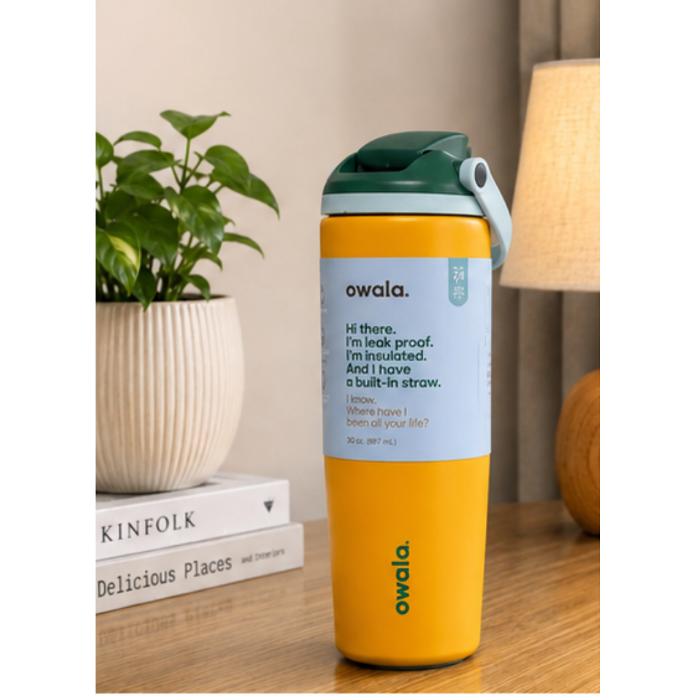 A yellow Owala water bottle with a dark green lid and a light blue wrap label, standing on a wooden table next to a potted plant and a lamp.