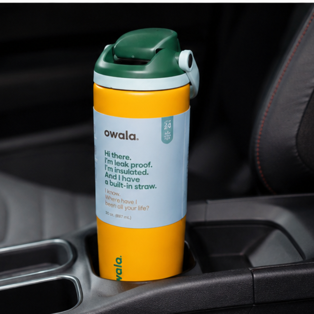 An Owala insulated tumbler with a yellow body, green lid, and light blue label, resting in a car cup holder.