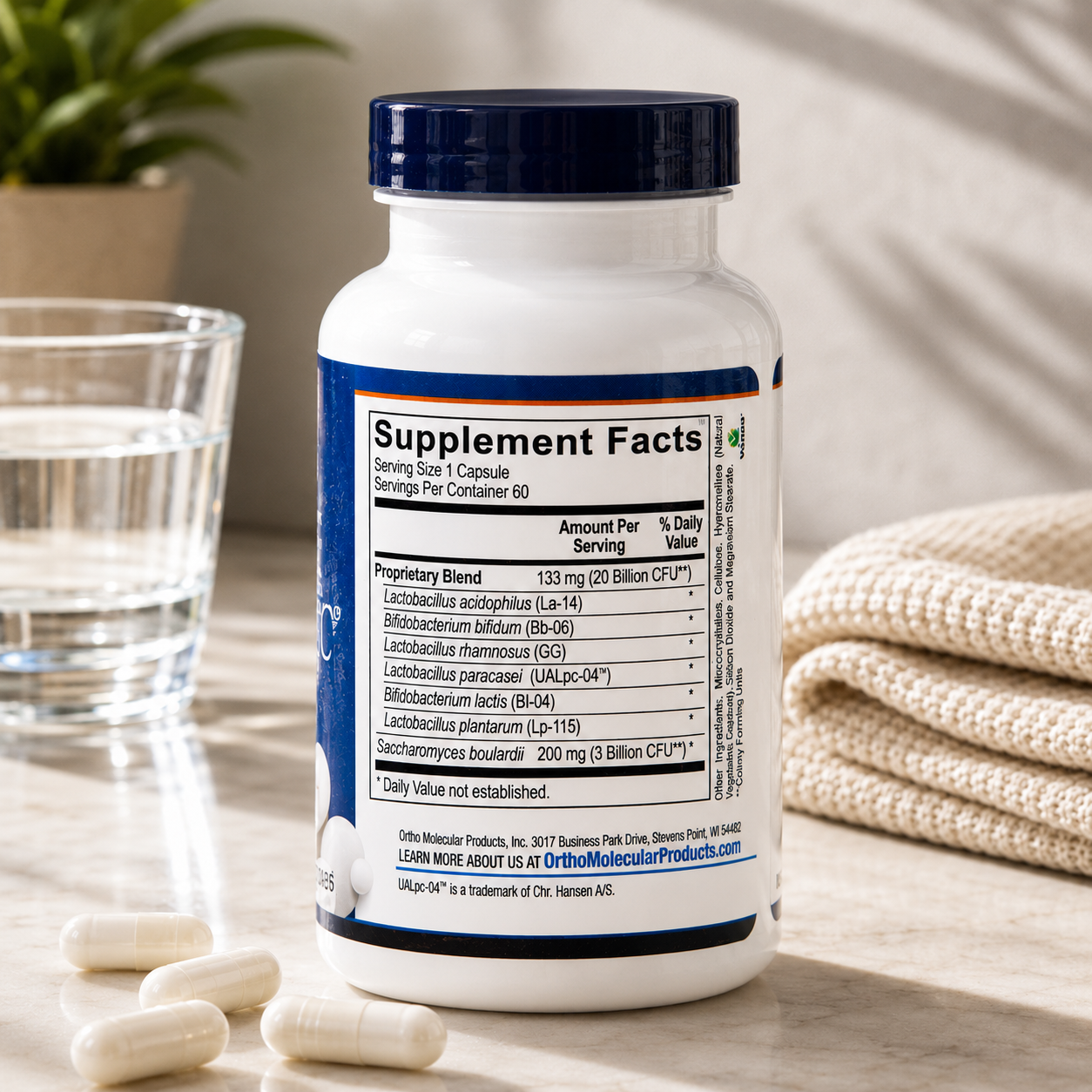A white supplement bottle with a dark blue cap stands on a tabletop, showing the Supplement Facts label. Several white capsules are scattered around it, with a glass of water on the left and a folded cream knit blanket on the right.