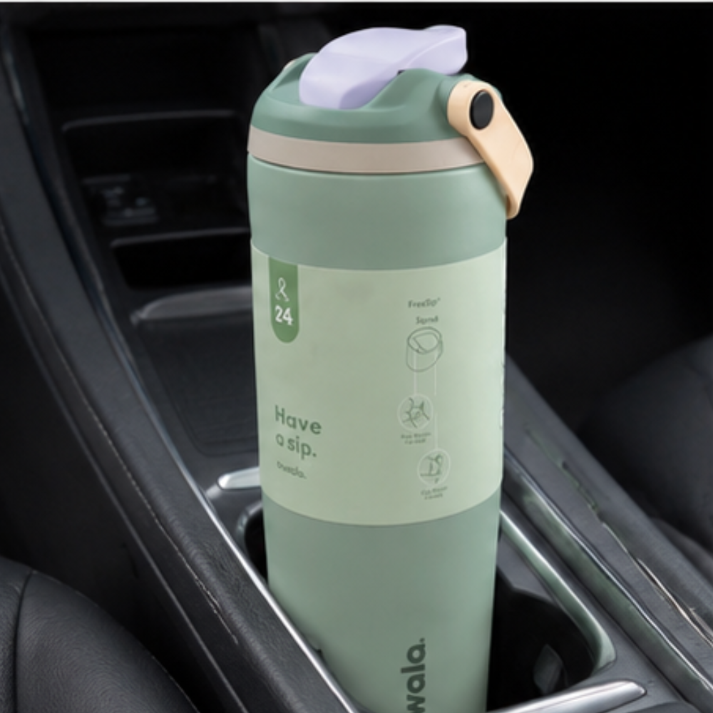 A mint green insulated water bottle with a light purple flip-top lid and beige carrying loop, placed in a car cup holder. The bottle has a white paper sleeve with product details and the brand name 'wala.